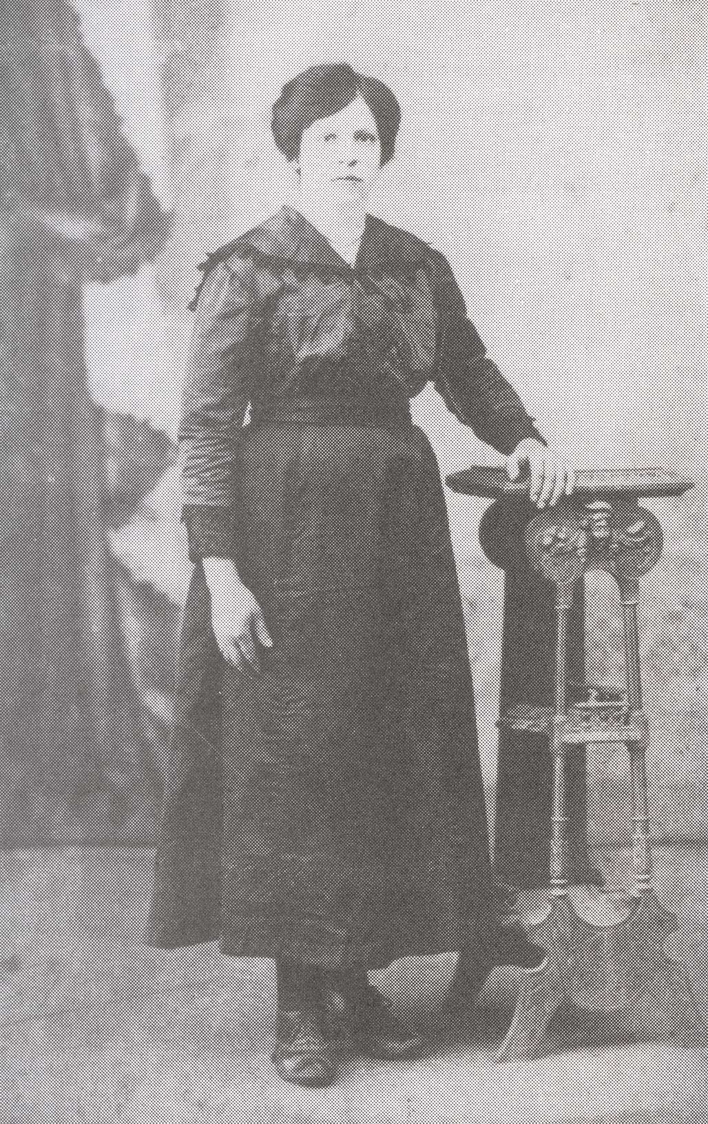 Very old image of a woman in a black dress, standing with her hand resting on a shelf.