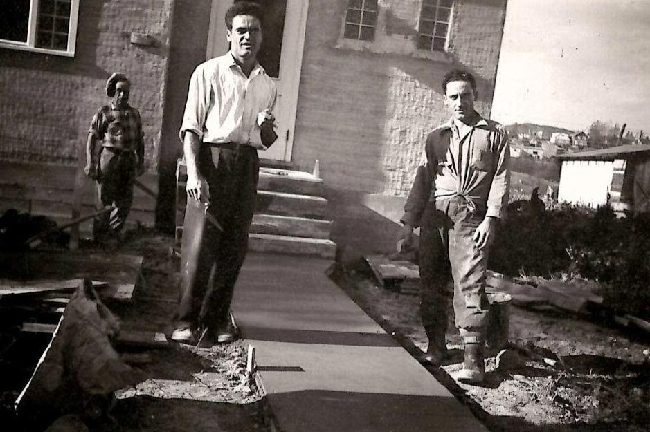 Black and white photo of three men laboring on cement walk outside house.
