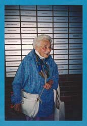 Grace as an elderly woman, standing in front of wall of plaques at Pier 21.
