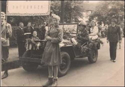 A small parade with a military jeep following two women holding up a banner.