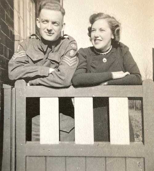 Black and white photo of a man in uniform and well-dressed young woman. They are both leaning on the wooden fence and standing with crossed arms.