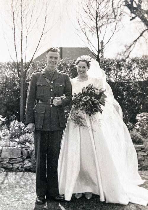 A young bride wearing a wedding dress and holding flowers stands next to a young man wearing a uniform.