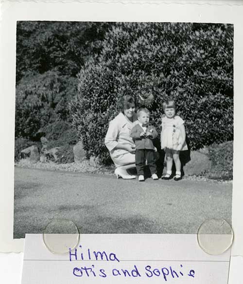 Woman kneeling next to small boy and girl, in front of a large hedge.