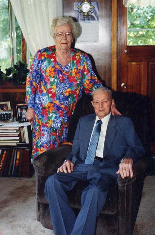An elderly man is seated and an elderly woman is standing next to him.