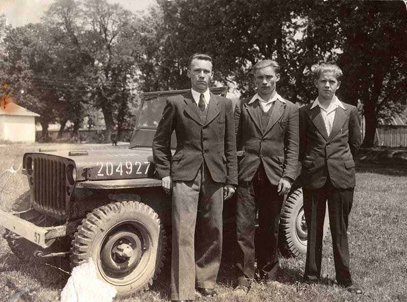 Three young men in dark suits stand in front of an old jeep.