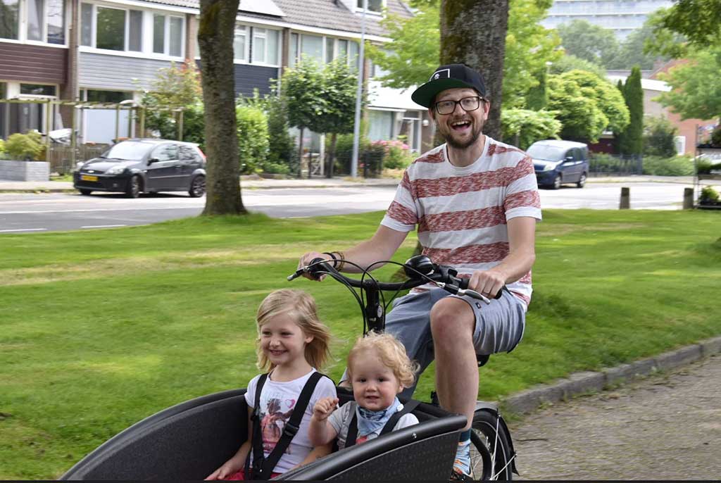 Young man rides a bicycle with two small children in the bucket seat.