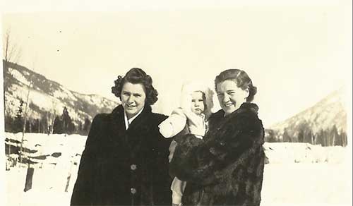 A young couple in winter clothing holding a baby as they stand in front of snowy hills.