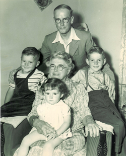 Older woman seated in armchair with three children around her and man standing.
