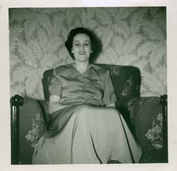 Joan in blouse and skirt, sitting on the sofa of a living room. 