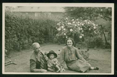 Young Joan wearing a hat, sitting in the grass of a garden with her parents. 