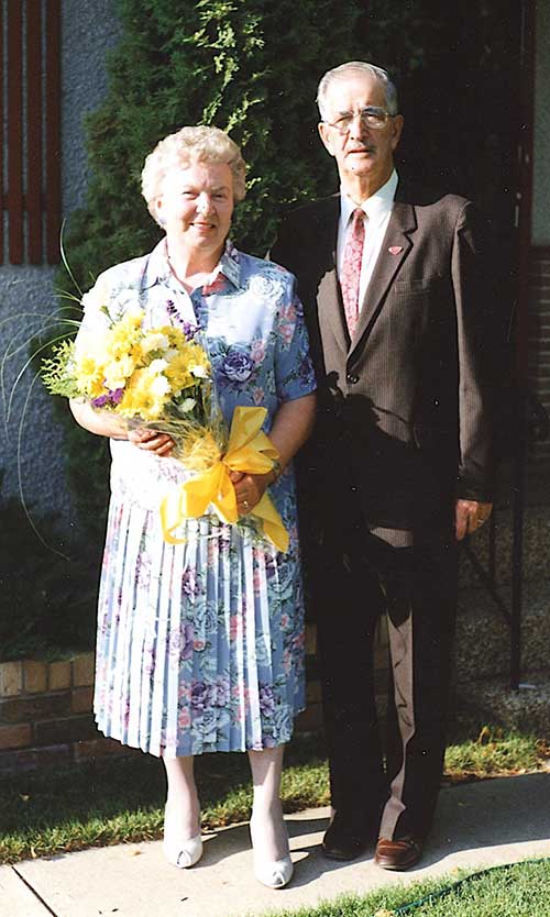 An elderly man and woman stand in front of their home, the woman holds a large yellow bouquet.