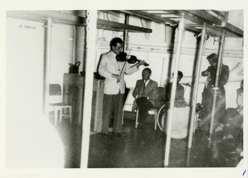 Old black and white image of man playing a violin on a ship while other people listen to him.