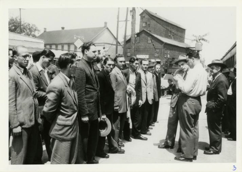 Two men stand facing a row of men as they read from a paper.