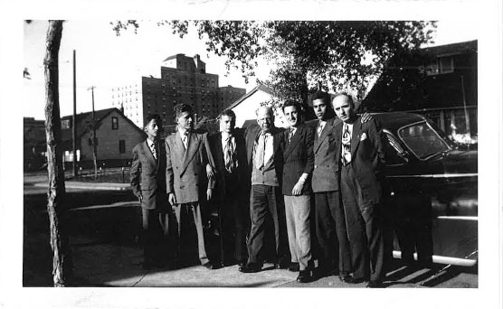 A group of men stand around a big black car.