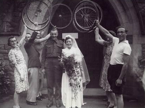 Young beautiful lady wearing wedding dress holding flowers. Standing with handsome man people standing around them holding cycle tires.
