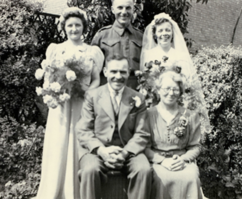Two young beautiful ladies wearing wedding dress holding flowers standing with handsome man and old couple sitting in front of them.