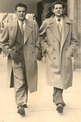 Two young men in suits and overcoats, walking on the sidewalk. 