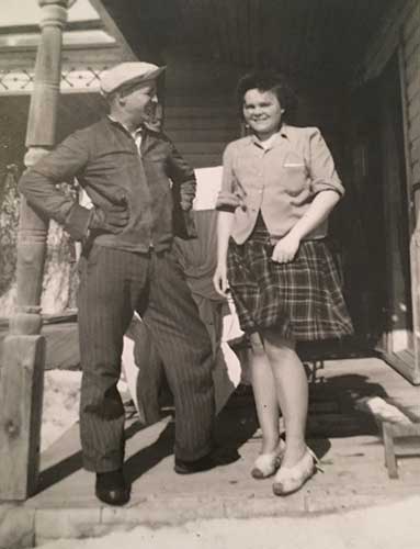 A man in cap and gloves is laughing and looking at the woman next to him, she is smiling and looking at the camera.