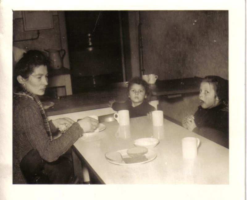 A young woman with two children sitting around the  dinner table, which has three cups and bread on it.