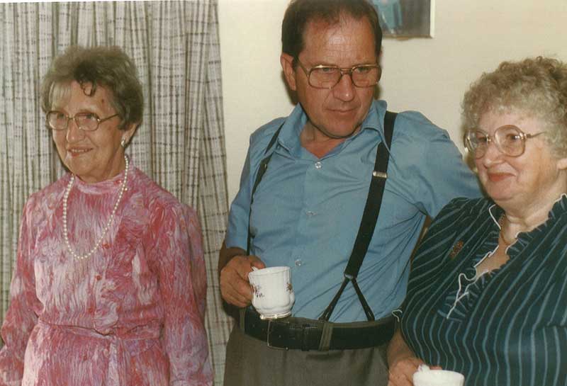 A man in suspenders holds a tea cup and two women stand on either side of him.