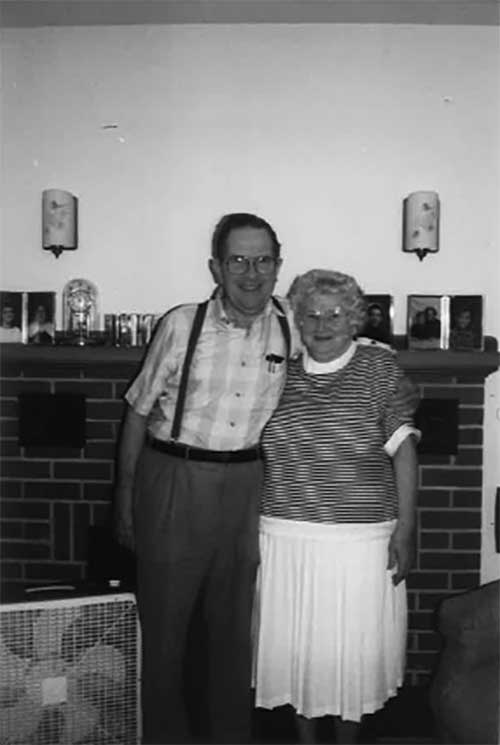 A man and woman are standing in front of a fireplace, there is a small fan next to them.
