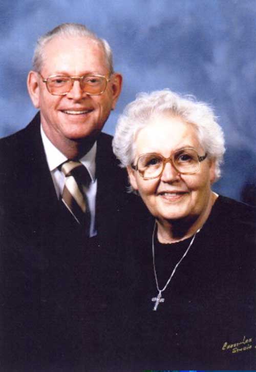 A middle-aged couple with grey hair stand wearing black and the woman is wearing a cross.