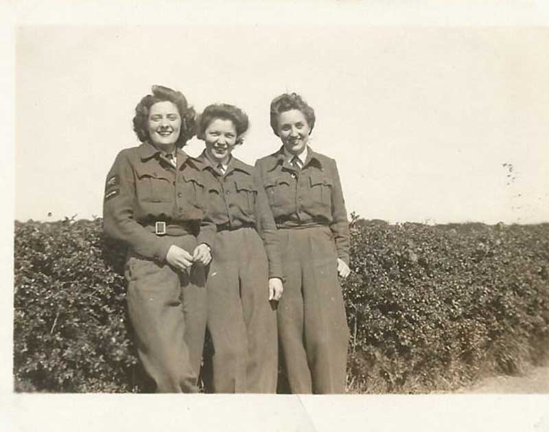 Three women stand in front of a hedge, they are all wearing military uniforms.
