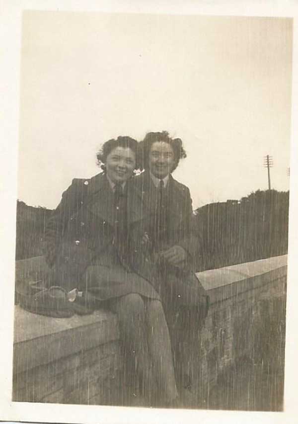 Two smiling women in military clothing are seated on a brick wall.