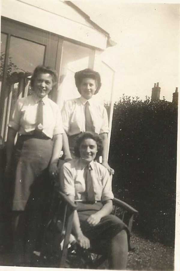 Three women are pictured, one seated and two standing behind her. They are all wearing white shirts and dark ties.