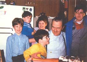 Older Leland, seated in kitchen surrounded by 5 grandchildren.