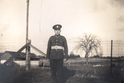 Young William in uniform, standing in front of a barbed-wire fenc.