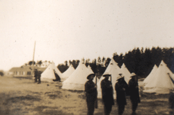 Four young men in military dress, standing at attention in front of tents.