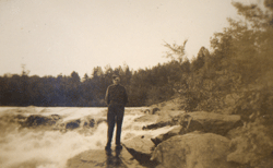 Young man in military uniform, standing on a rock next to flowing river.