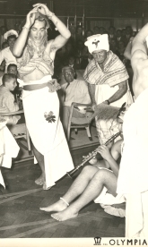 Young man sitting on the floor, playing the flute while costumed people dance around him.