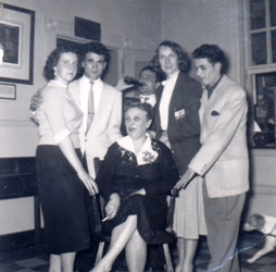 Woman in black dress seated, with five young adults standing around her.
