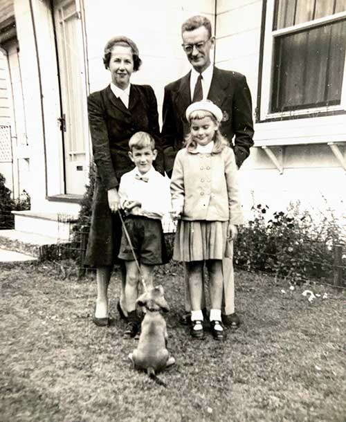 A young man and woman with two children and a dog are standing in front of a house.