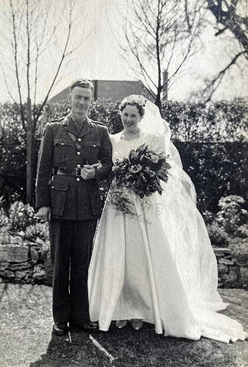 A young woman is wearing a wedding dress and holding flowers, while standing next to a young man in military uniform.