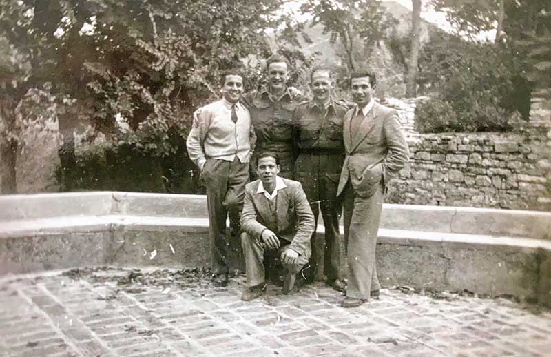 Group of five people standing and sitting in front of trees, having their photo taken.
