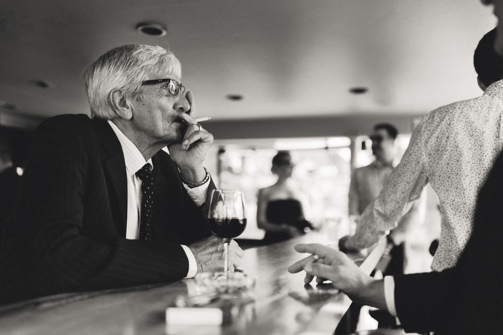 A black and white photo of an Italian man in a suit smoking a cigarette at a bar with a glass of wine.