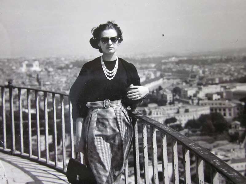 A well-dressed young woman stands against a railing which overlooks a beautiful city.