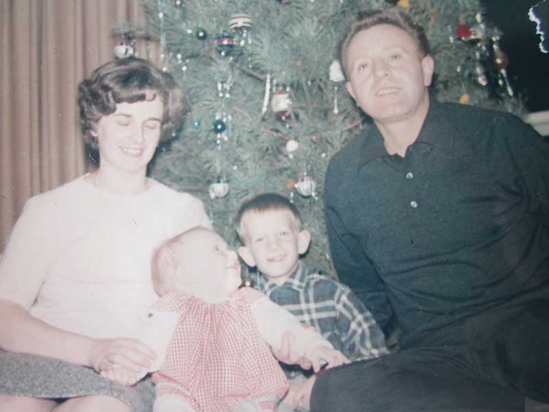 A man and woman sit in front of a Christmas tree, with two laughing children between.
