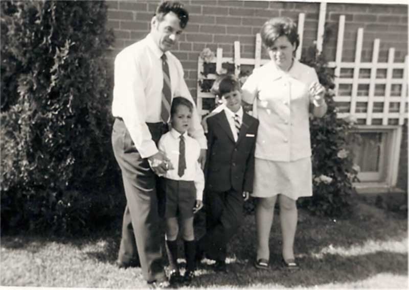 A young family pose outside their home; father and mother and two young boys dressed in school uniforms.