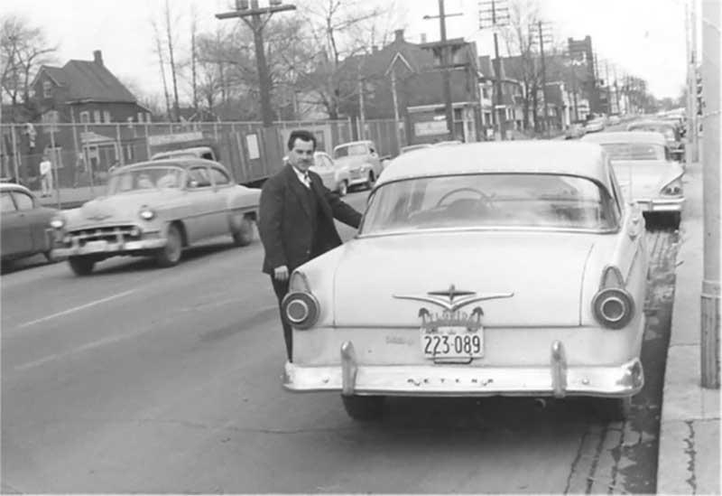 A young man stands beside his car as he looks at the camera.