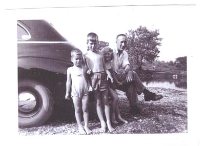 Young George and three children sitting on the rear bumper of a car.