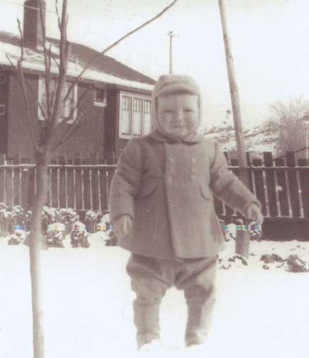 A baby standing on snow in front of house.