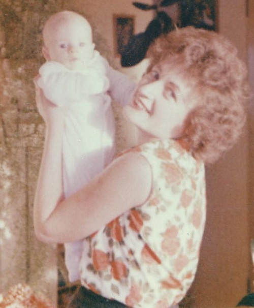 Young woman with red hair holds up a beautiful baby dressed in white.