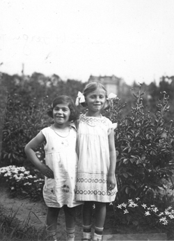 Two young girls in a garden, wearing white summer dresses and ribbons in their hair.