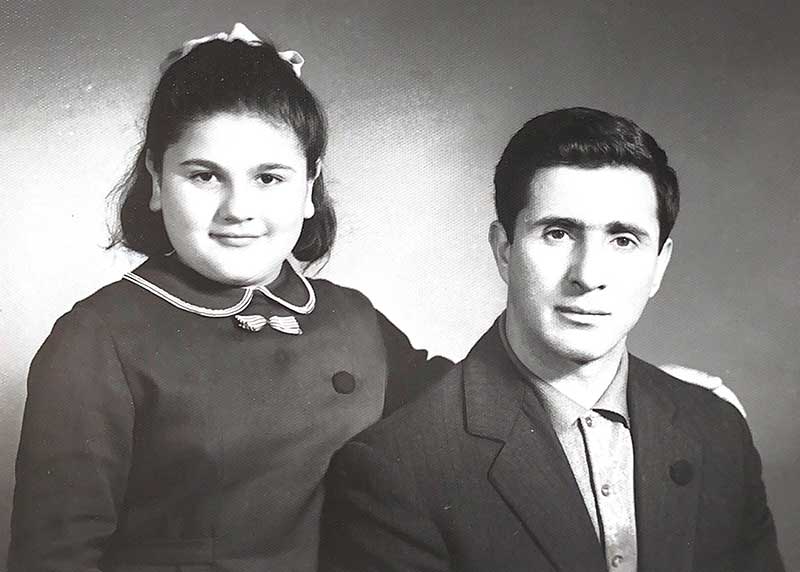 A young girl with a bow in her hair, stands next to a seated gentleman.