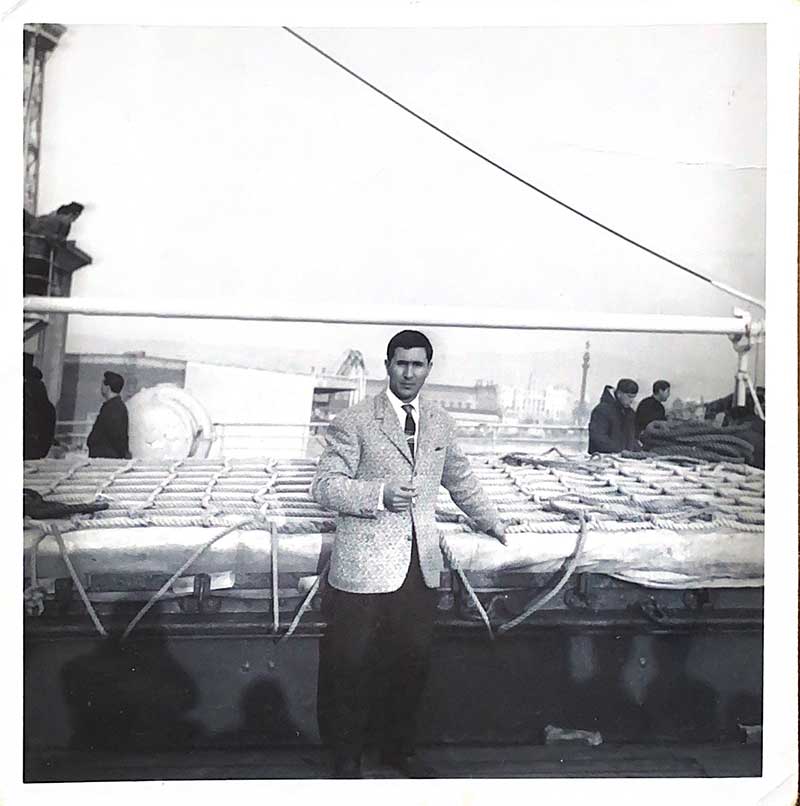 A man in a suit is standing on the deck of a ship, he is smoking a cigarette.