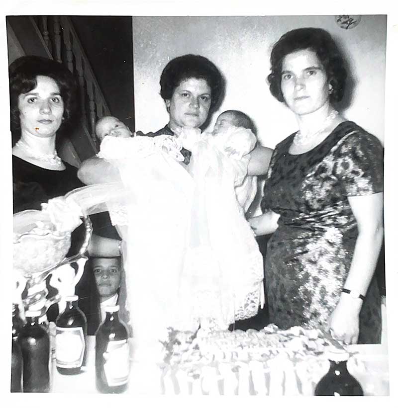 A woman holds twin babies in christening gowns, two women are standing on either side of her.